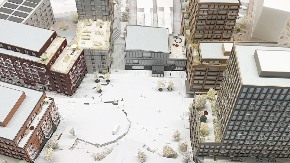 Flerbostadshus runt ett torg, sett uppifrån. Husen har olika höjder, storlekar och färger på fasaderna. På några av taken ses planteringar och terrasser. På torget finns planterade träd, öppen yta med trappavsatser, sittplatser. Illustration